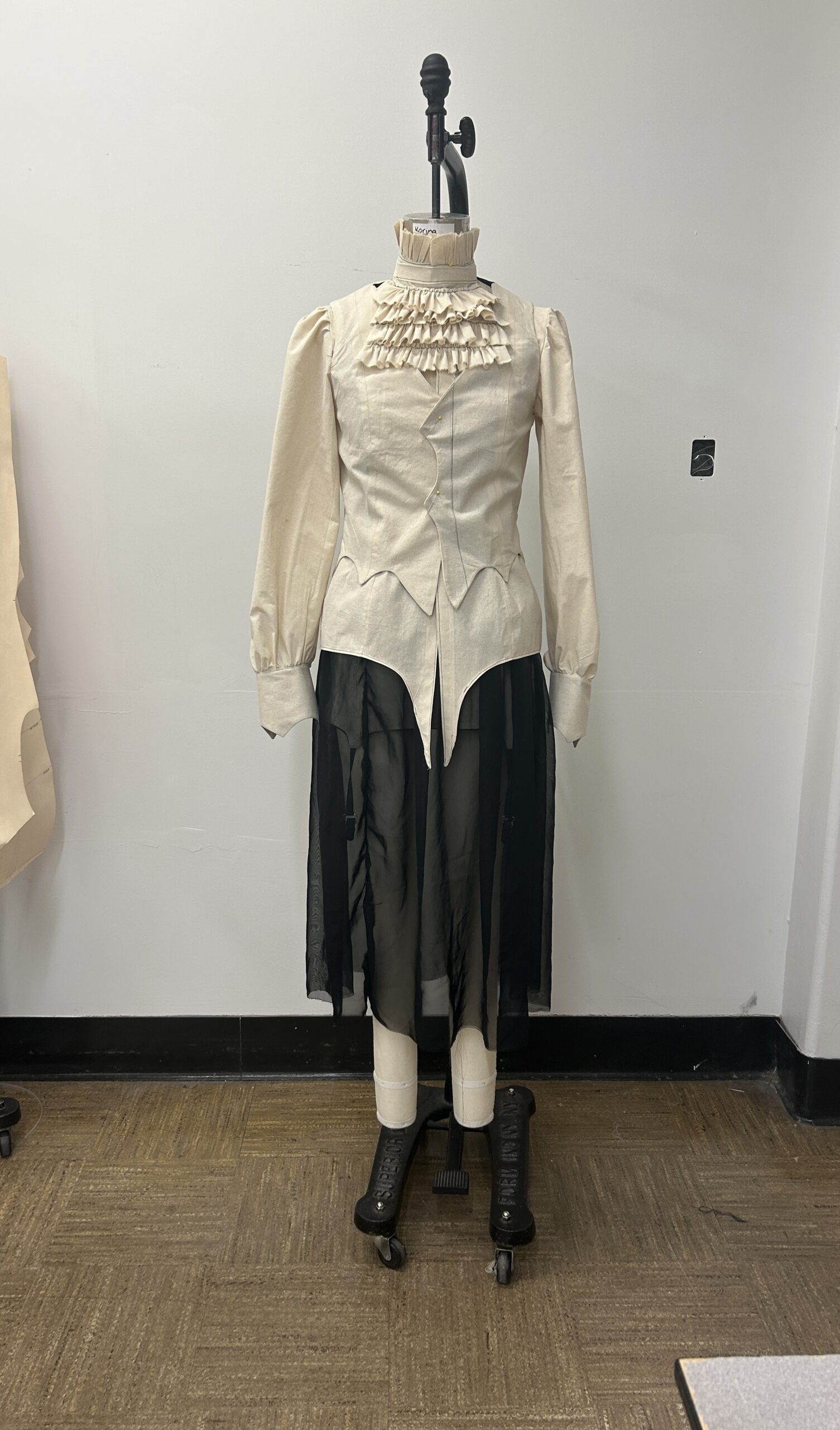 Muslin samples of one look from the collection on a dress form, featuring a puff sleeve shirt, waistcoat, and mini skirt with sheer over-skirt.